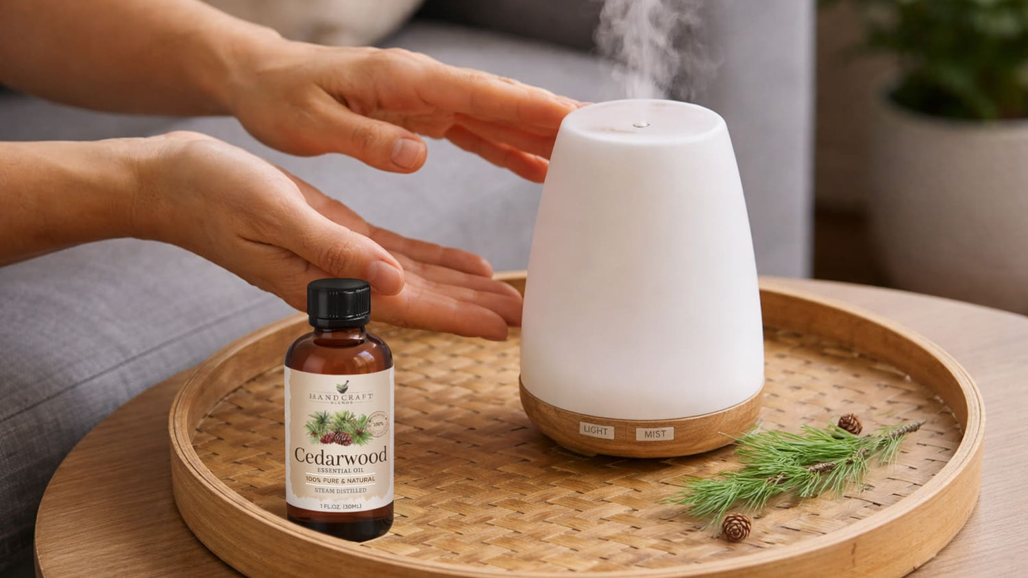 Cedarwood essential oil bottle with hands near an aromatherapy diffuser on a wooden tray