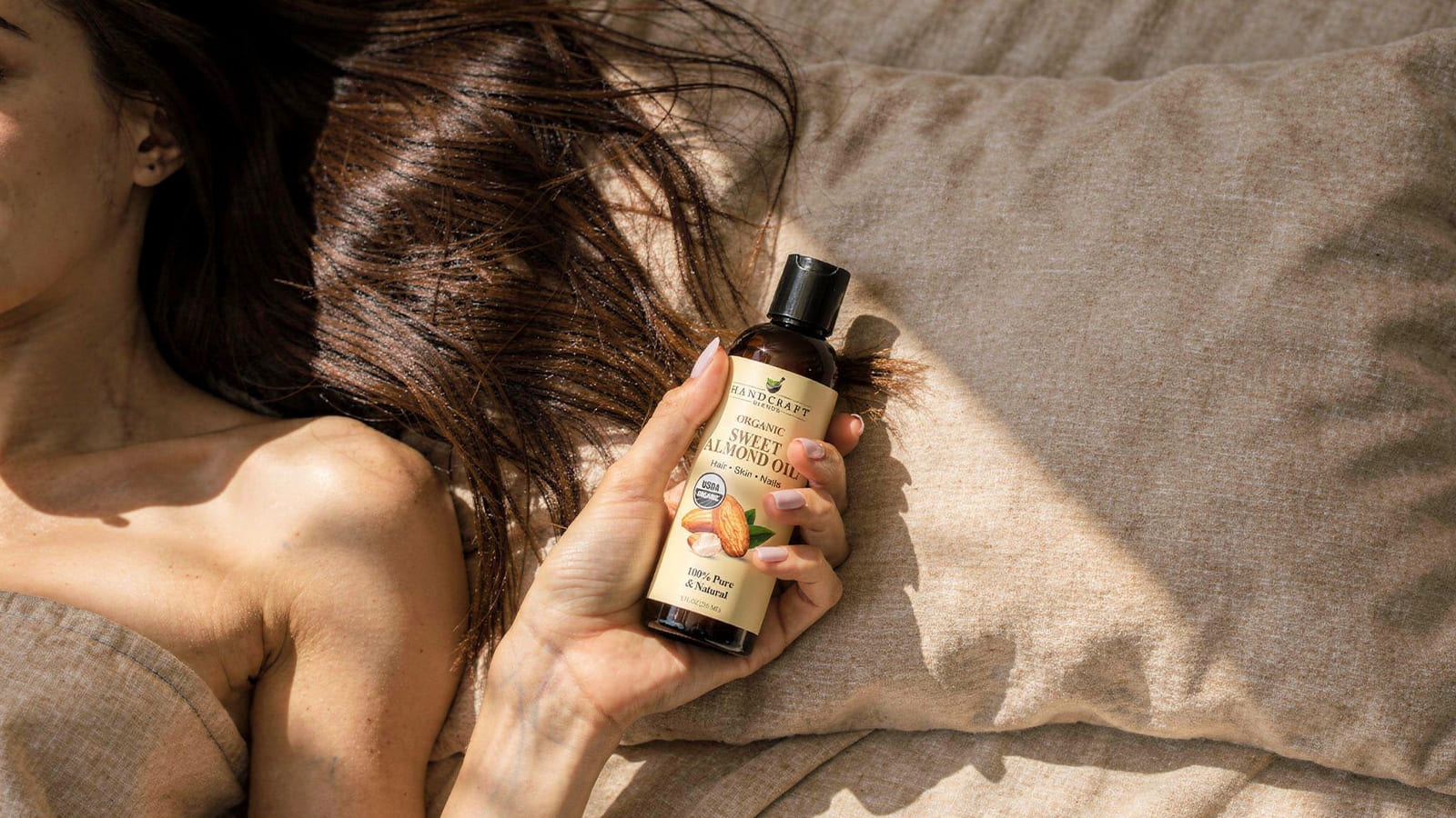 Woman holding bottle of organic sweet almond oil on a beige bed for skin and hair care