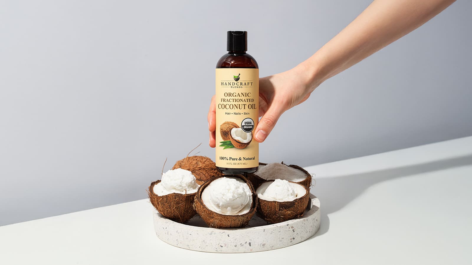 Hand holding bottle of organic fractionated coconut oil above halved coconuts on tray
