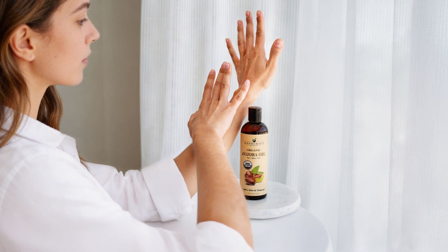 Woman applying organic jojoba oil to hands, bottle on table, natural skincare
