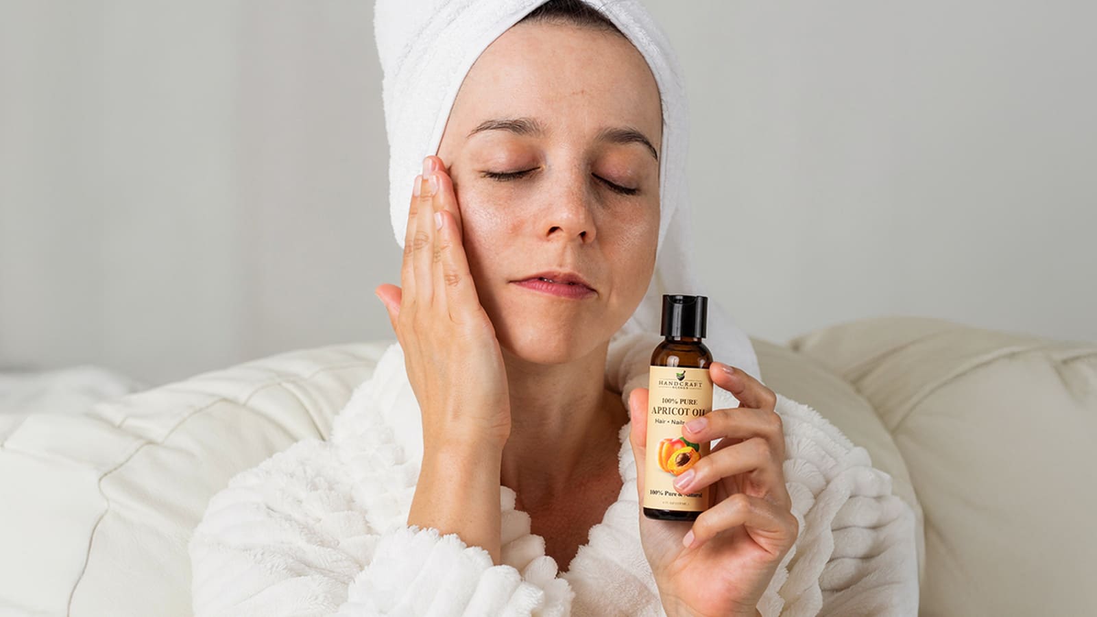 Woman applying apricot oil to face for skincare, holding bottle, spa robe and towel