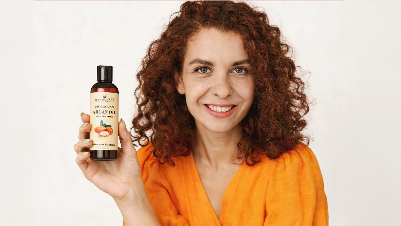 Smiling woman with curly hair in orange shirt holding Moroccan argan oil bottle for hair and skin