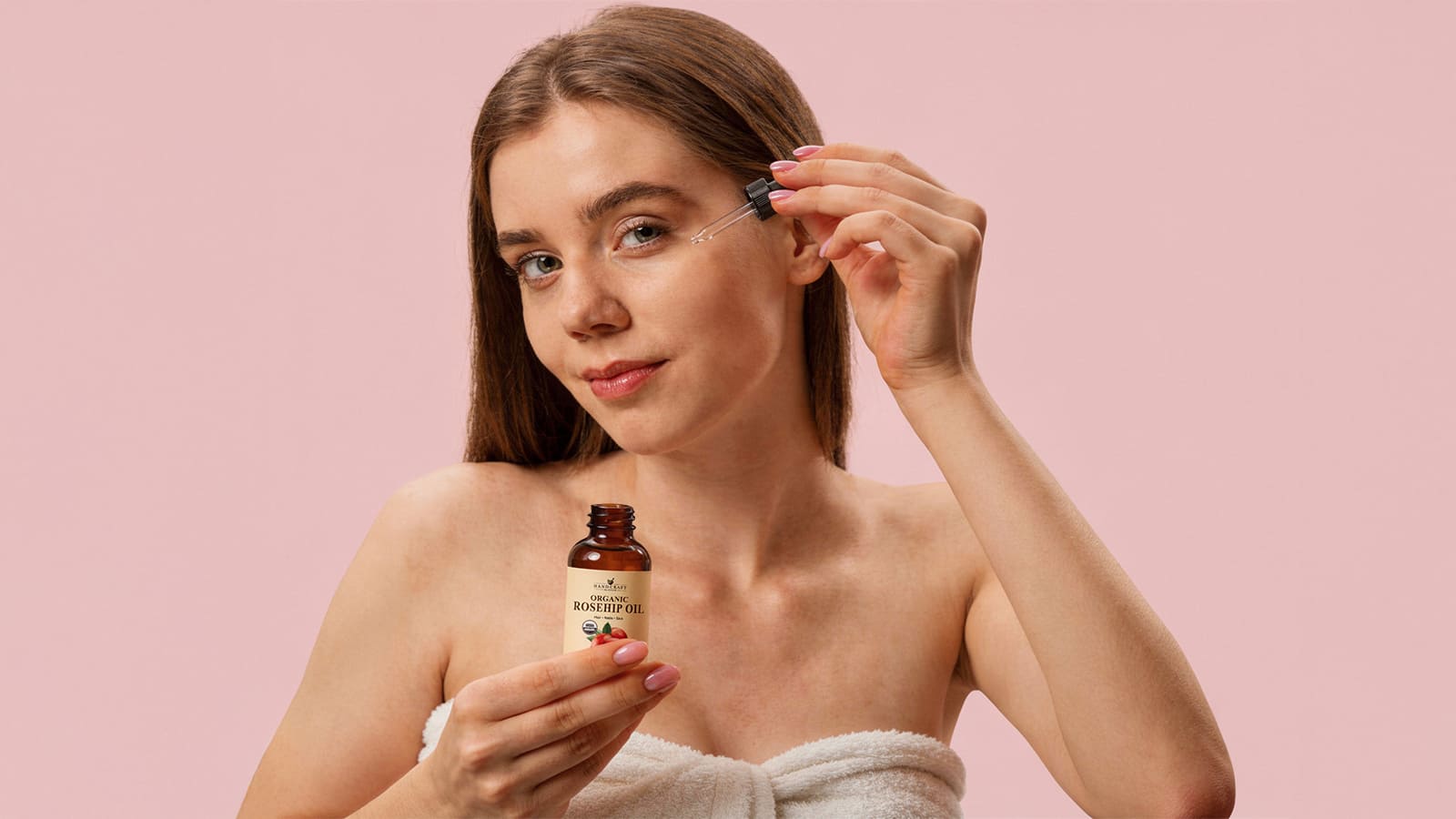 Woman applying organic rosehip oil to face with dropper on pink background