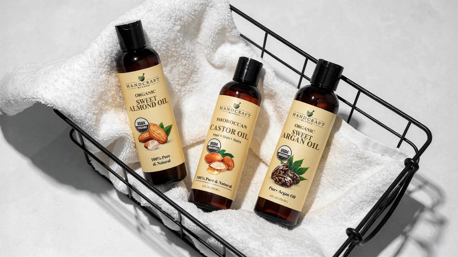 Three bottles of organic carrier oils—sweet almond, castor, and argan oil—in a basket on a white towel.