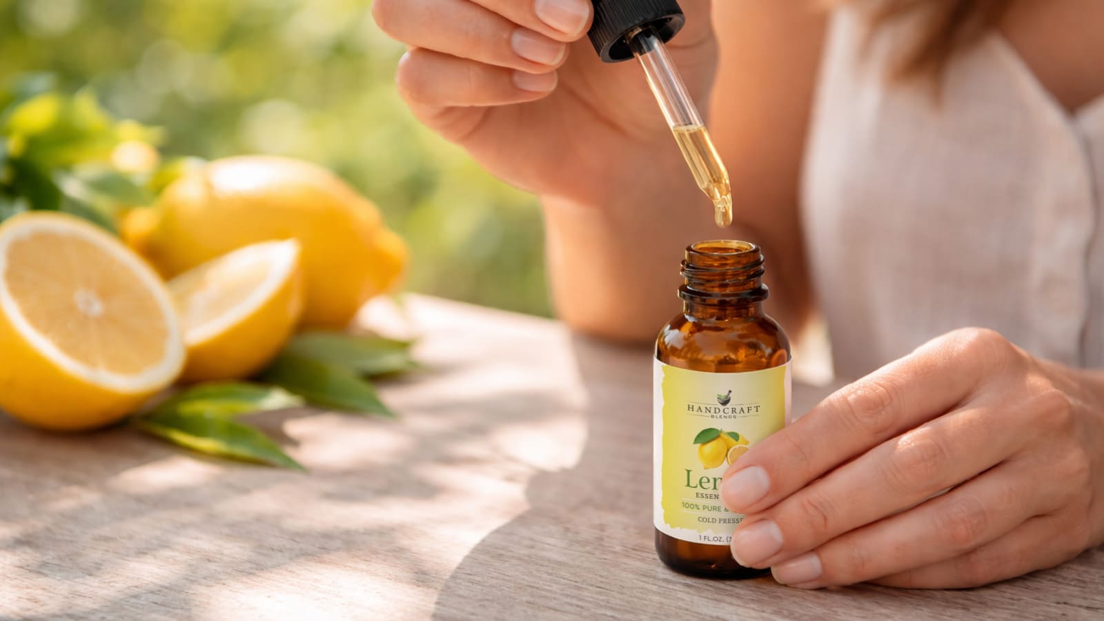 Hand using dropper with lemon essential oil bottle, fresh lemons and leaves on table