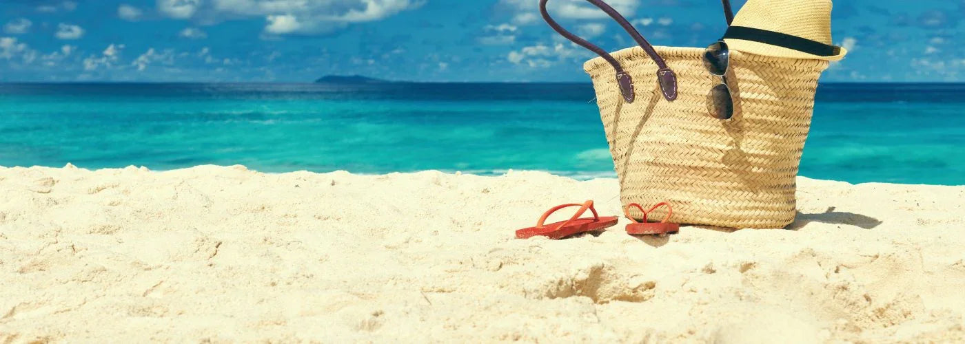Woven beach bag, straw hat, flip-flops, and sunglasses on sandy shore by blue ocean