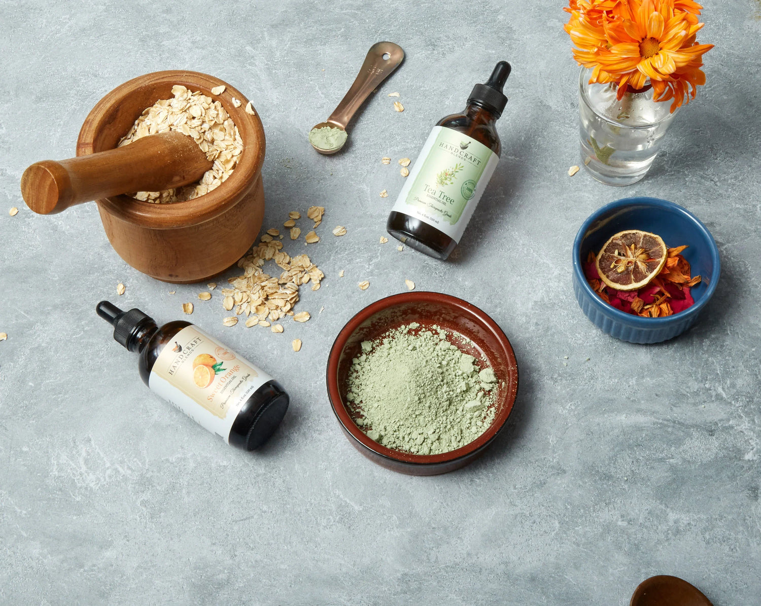 Handcraft Blends essential oils, oats, green clay, dried botanicals, and flowers on a gray surface